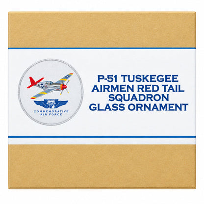 A small rectangular product box for a P-51 Tuskegee Airmen Red Tail Squadron glass ornament, photographed on a dark reflective surface. The box is tan with a white center label showing a red, white, and blue airplane emblem and blue text reading 'P-51 TUSKEGEE AIRMEN RED TAIL SQUADRON GLASS ORNAMENT'.
